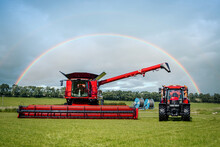 Brand New Modern Clean Combine Harvester And Shiny Red Agricultural Tractor In Rural Farm Field On Summers Day With Beautiful Colourful Rainbow In The Sky Behind