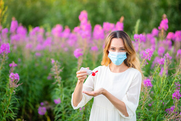 A girl in a medical mask wraps her hands with a sanitizer while standing in a field against a background of flowers