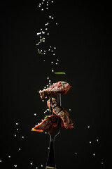 Canvas Print - Grilled beef steak on a fork adding rosemary and sea salt in a freeze motion on black background. vertical image, place for text.