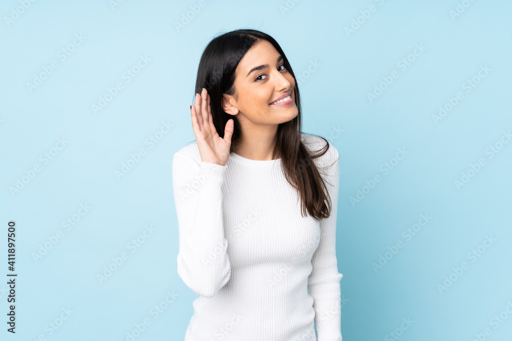 Young caucasian woman isolated on blue background listening to something by putting hand on the ear - obrazy, fototapety, plakaty 