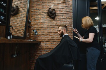 Stylish man sitting barber shop Hairstylist Hairdresser Woman cutting his hair Portrait handsome happy young bearded caucasian guy getting trendy haircut Attractive barber girl working serving client