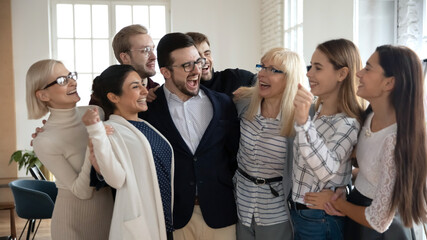 Group of happy workers of different generations hugging like family together, laughing. Excited diverse business team cheer celebrating success. Corporate achievement, win, teamwork, community concept