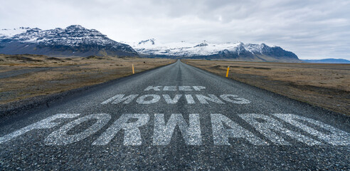 Keep moving forward text quote written on asphalt road leading towards infinity and mountain scenery in the background.