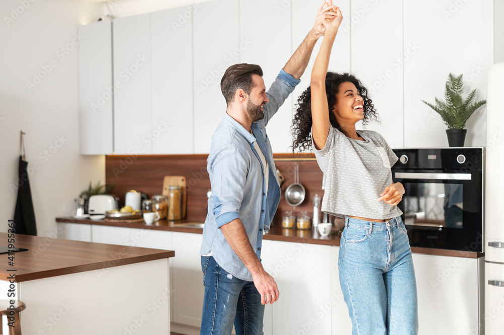 Handsome white man and attractive African American young woman is cooking and having fun together while cooking on the kitchen, romantic interracial couple is dancing while preparing lunch or dinner - obrazy, fototapety, plakaty 