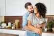 © Vadim Pastuh - Smiling man hugging from behind charming African American woman, two people standing and joyfully looking at each other. Young international couple happily spending time in cozy modern kitchen at home