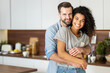 © Vadim Pastuh - Smiling man hugging from behind charming African American woman, two people standing and joyfully looking at camera. Young international couple happily spending time in cozy modern kitchen at home.