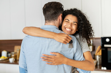 Delighted pleasant young African American woman hugging her boyfriend and feeling thankful for buying a new house, holding keys, and smiling, interracial couple homeowners celebrating the relocation