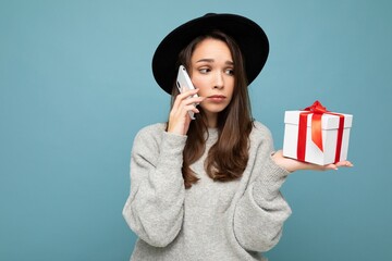 Photo of beautiful upset young brunette woman isolated over blue background wall wearing black hat and grey sweater holding gift box talking on mobile phone and looking to the side