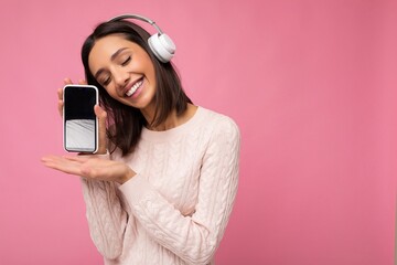 Attractive positive smiling young woman wearing stylish casual outfit isolated on colourful background wall holding and showing mobile phone with empty screen for cutout wearing white bluetooth