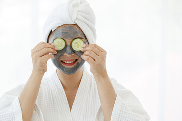 A cute Asian woman wearing a spa dress and covered her head with a white towel and added treatment cream on her face holding and playing with pieces of cucumber with happiness manner