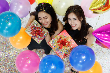 Two young and beautiful sexy Asian girls with colorful gift box dancing and playing with fun and happiness in a party with the bright color balloon in the background. Taken from a top-down angle.