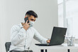 © fotofabrika - Handsome man businessman in medical mask using his laptop while talking on his cellphone