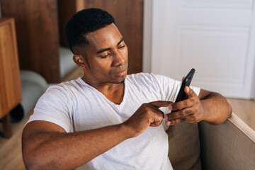 High-angle shot of focused African-American man in white T-shirt sitting at armchair in domestic room and using mobile phone. Handsome male browsing internet on cellphone.