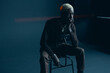 dark-skinned handsome guy with white hair and blue eyes sitting on a chair in a dark studio with a serious facial expression in brown clothes and silver accessories, he looks away