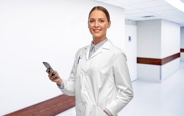 Canvas Print - medicine, profession and healthcare concept - happy smiling female doctor with stethoscope using smartphone over hospital corridor background