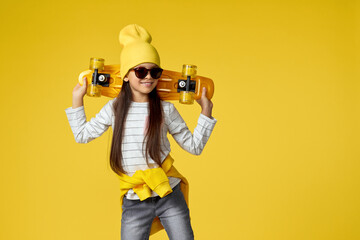 cute little child girl in hat and sunglasses posing with yellow skateboard on studio background. copy space