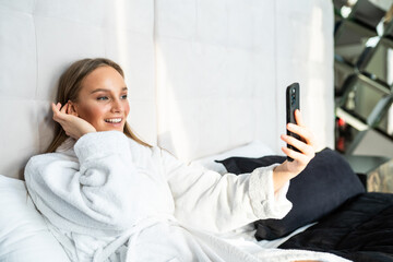 Wall Mural - Young beautiful woman with towel on a bed in a hotel room, dressed in a white terry bathrobe, taking selfie on her mobile phone, a slight smile on her face