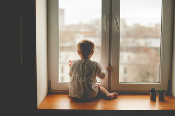 Cute caucasian infant toddler child sitting on the window with his back, baby safety and waiting, lifestyle and toning