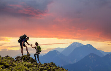 tourist couple helping each other to climb high rock in evening mountains at sunset.