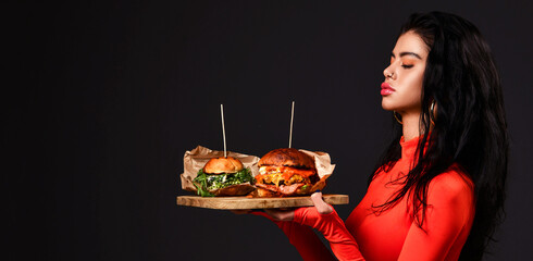 Wall Mural - Pretty brunette woman with closed eyes and pouty lips in red bodysuit holds two fresh big burger on wooden tray over black background with copy space. Side view. Banner. Fast food and dieting concept.