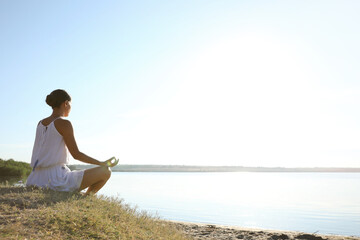 Wall Mural - Young woman meditating near river at sunset, space for text. Nature healing power