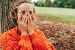 © iuricazac - Horizontal candid portrait of a happy young woman smiling, hiding her face with two hands, playing peek a boo game in the park. Female wearing an orange sweater has joyful expression outdoors.