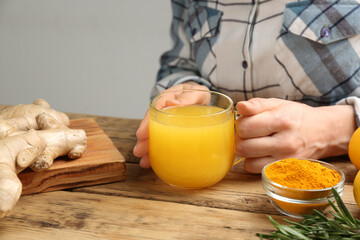 Woman holding cup of immunity boosting drink at wooden table with ingredients, closeup