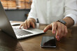 © New Africa - Freelancer taking smartphone while working on laptop at table indoors, closeup