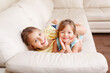 © anoushkatoronto - Two cute little Caucasian girls siblings playing at home. Adorable smiling children kids lying on a couch together. Authentic candid lifestyle domestic life moment. Happy friends sisters relationship.