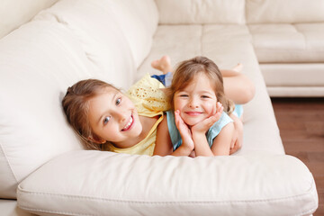 Two cute little Caucasian girls siblings playing at home. Adorable smiling children kids lying on a couch together. Authentic candid lifestyle domestic life moment. Happy friends sisters relationship.