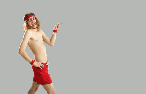 Comic skinny athlete laughing pointing finger at free space for text on gray background. Man in red shorts, glasses and a headband defiantly ridicules. Concept of funny sport, advertising and banner.