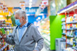 © luciano - Adult senior man with surgical mask due to coronavirus while shopping at the supermarket. White haired retiree with shopping cart, choosing a canned product. Consumerism concept