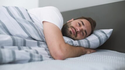 Canvas Print - Calm young bearded man wears casual blue t-shirt lying in bed sleep with folded hands under cheek rest relax spend time in bedroom lounge home in own room house wake up open eyes. Real estate concept