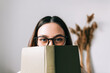 © nikkimeel - Portrait of young caucasian woman college student in eyeglasses hiding behind a book and looking at camera.