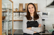 © Vadim Pastuh - Confident female business owner standing at the counter of a restaurant with arms crossed, bakery waitress in apron looks at the camera and smiles