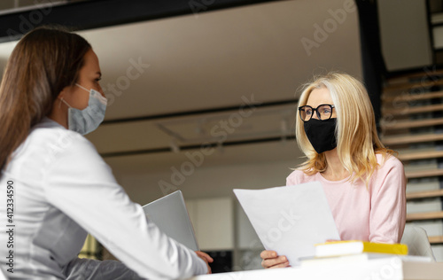 Mature blonde female hr manager sitting at the desk, wearing face mask, training young female worker intern, interviewing job seeker, checking cv, mentoring student. Social distance and safety concept