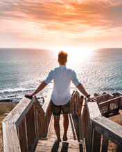 Back View Of A Young Man Standing On Wooden Stairs Leading To The Sea At Sunset