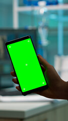 Close up of biochemist holding and looking at phone with chroma key display standing in biological lab while nurse in white coat bringing blood sample. Scientist using phone with mockup, green screen