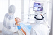 © DC Studio - Little girl in ppe looking at her mother in the course of dental examination. Stomatolog in protectie suit for coroanvirus as safety precaution looking at digital child teeth x-ray during consultation