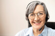 © Vadim Pastuh - Close-up of the face of a charming beaming elegant elderly lady, wearing headset, glasses, smiling and looking at the camera. Portrait of a senior customer service representative sitting in the office