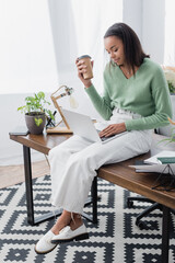 Canvas Print - smiling african american interior designer sitting on desk with paper cup and using laptop