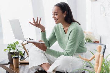 Canvas Print - cheerful african american freelancer sitting on desk and gesturing during video chat on laptop, blurred foreground