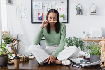 Canvas Print - cheerful african american architect holding pencil near nose while sitting on desk with crossed legs