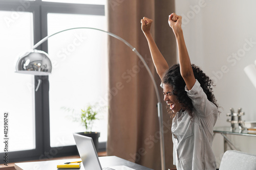 Happy excited African American businesswoman celebrating success, competition winner raising hands high, expressing joy from achieving goals, sitting at the desk with an open laptop in the office