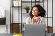 © Vadim Pastuh - Smiling young joyful African American female freelancer enjoying morning coffee while working on the laptop, looking away sitting at the modern desk at home, happy businesswoman having a coffee break