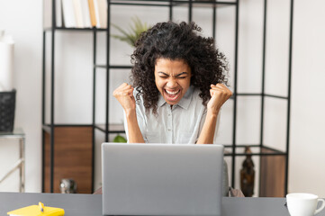 Happy cheerful African American woman with raised fists celebrating success, making yes gesture, looking at the laptop screen, passed exam test with a high score, got promoted, victory winner concept