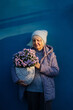 © igishevamaria - Smiling aged woman in casual warm wear holding pot with blooming pink flowers  while standing against blue wall. Positive elderly woman carrying potted flowers while representing  gardening concept.