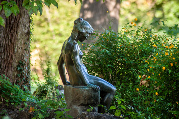 Nymph in woods - Garden statue of nude woman sitting on stump hidden among the trees and flowers in a public park.