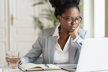 Focused Young African American Businesswoman Or Student In Blazer Wear Glasses Working At Laptop At Home Office, Holding Glass Of Water, Looking At Screen Computer, Reading Interesting Article Online.