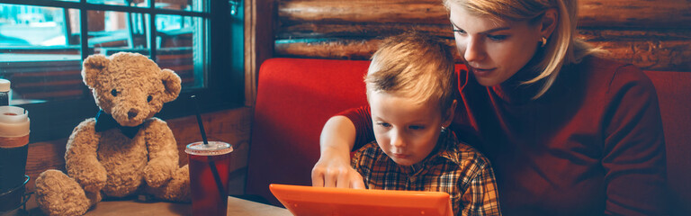 Caucasian mother and boy son choosing food meal from digital tablet touch pad menu at restaurant. Mom and kid playing on digital children gadget. Family is going out. Web banner header.